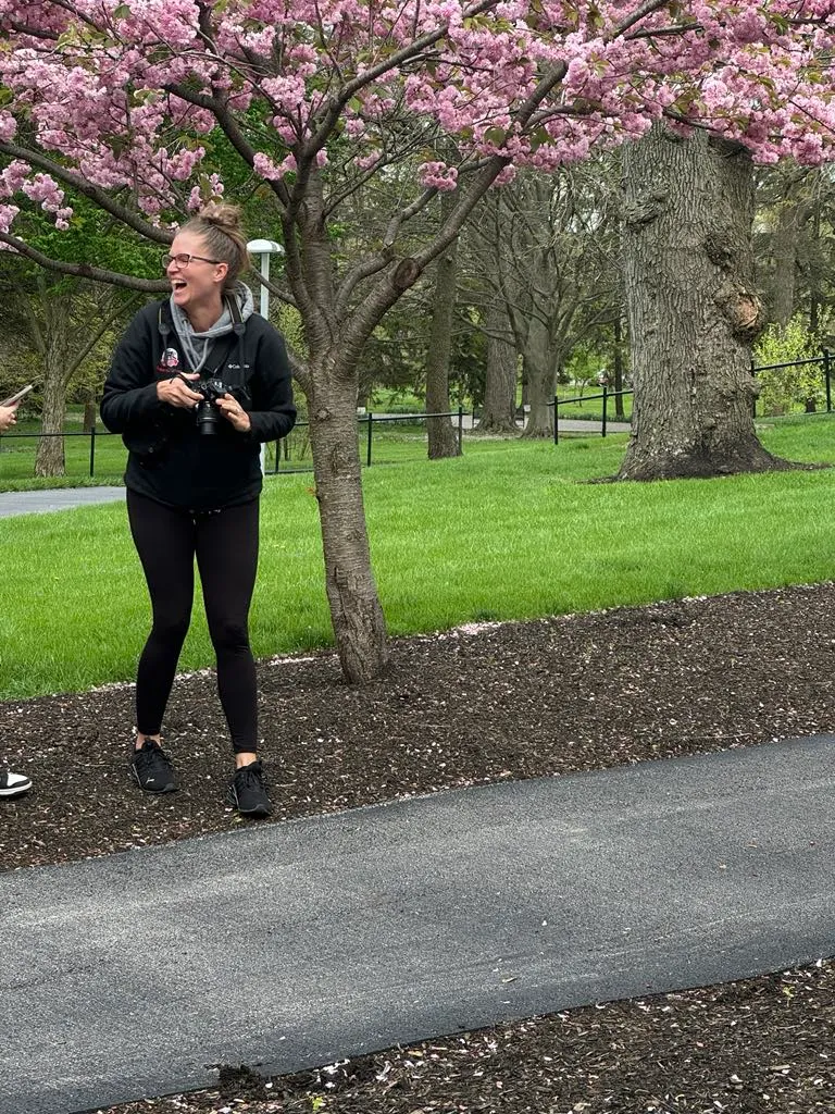 Photographer Betsy laughing while holding DSLR camera under pink cherry blossom tree in park